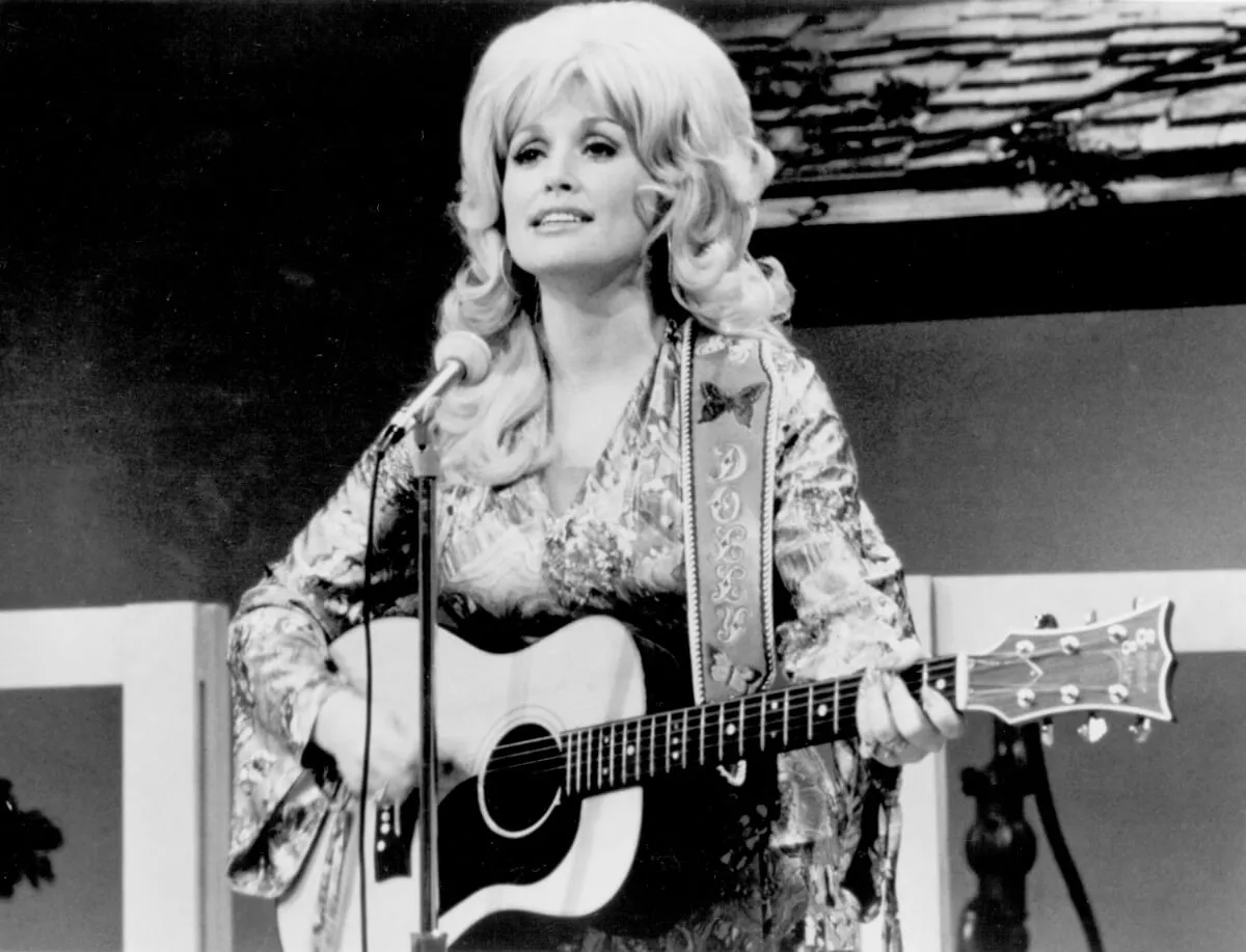 A black and white picture of Dolly Parton playing the acoustic guitar in front of a microphone.