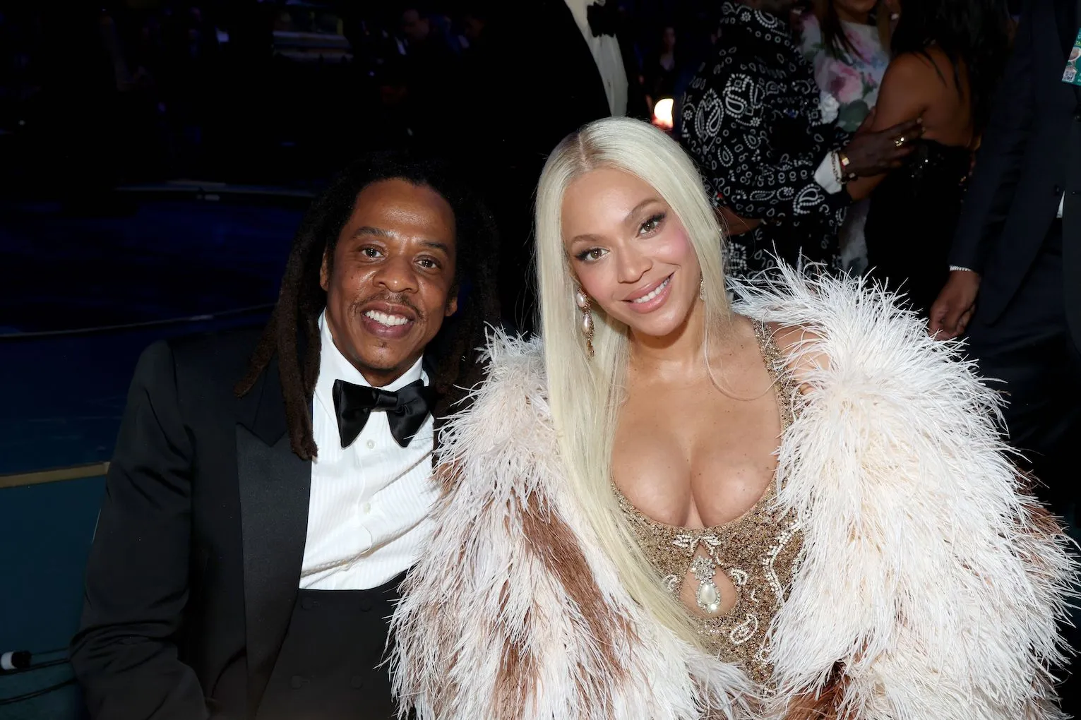 Jay-Z and Beyoncé sitting together and smiling at the 2025 Grammy Awards. Jay-Z is wearing a tuxedo. Beyoncé is wearing a fur coat over a gold dress. Her hair is long and blonde.