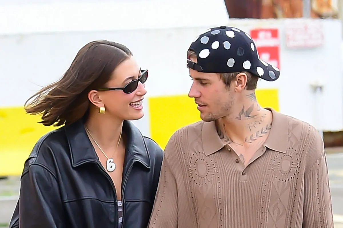 Hailey Bieber wears sunglasses and a leather jacket. She walks with Justin Bieber, who wears a tan shirt and a backwards hat.