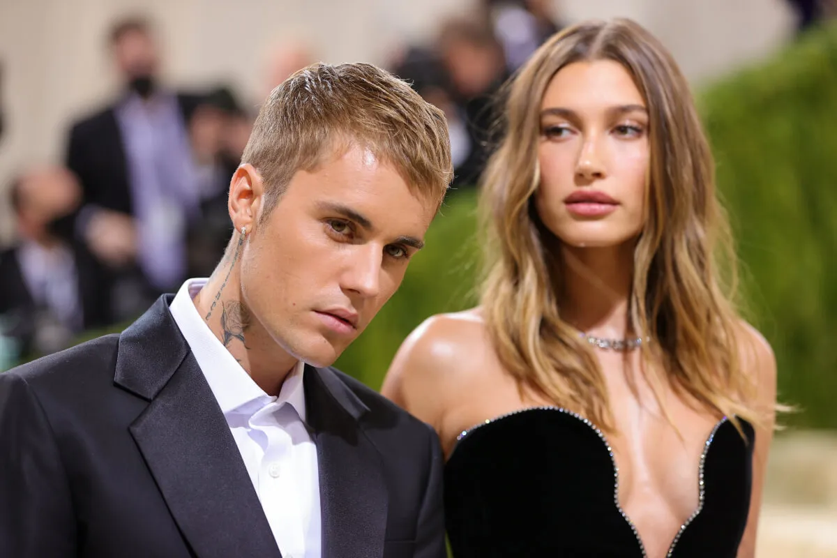 Hailey Bieber wears a strapless black dress and stands behind Justin Bieber. He wears a suit.