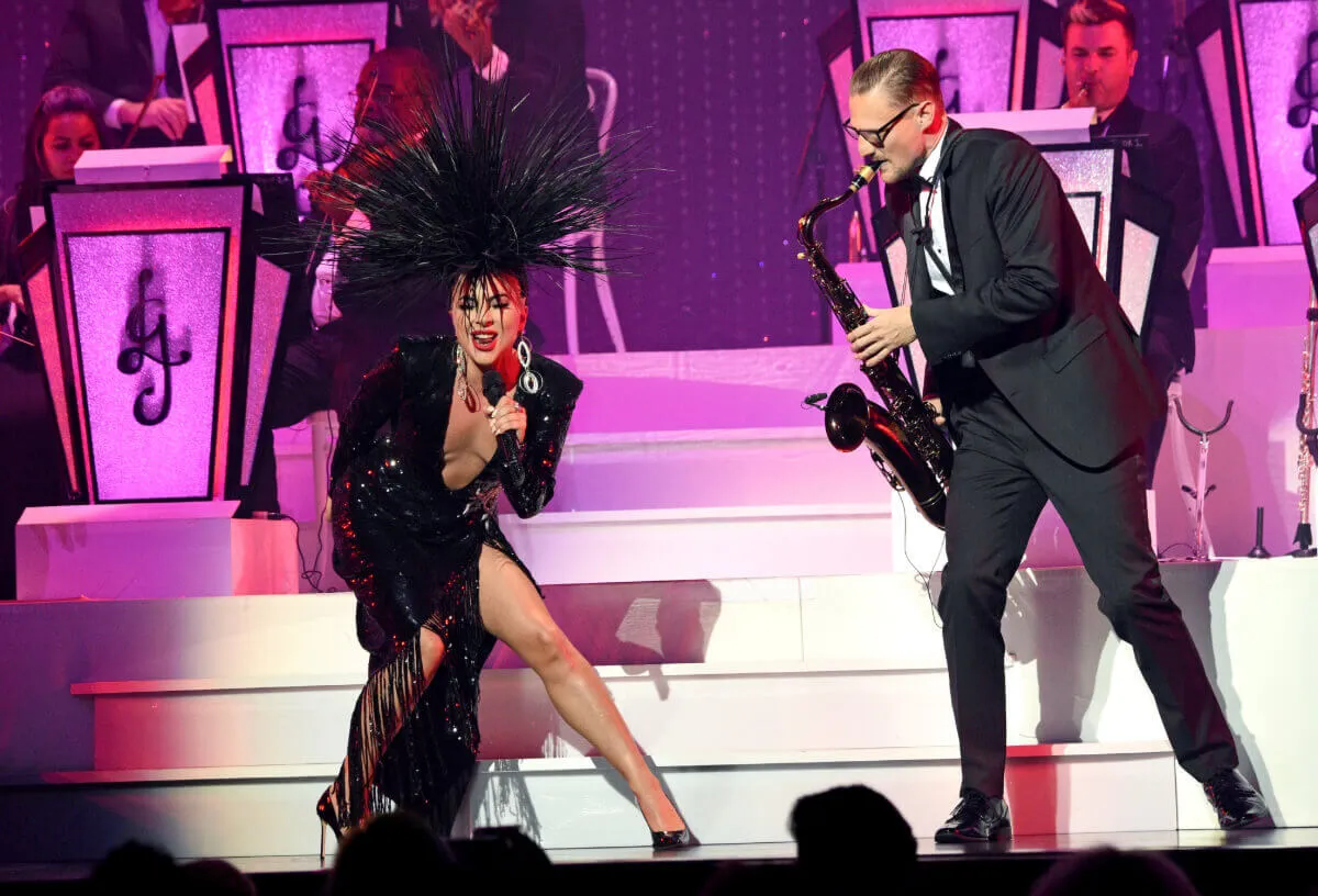 Lady Gaga wears a headdress and sings on stage during her Las Vegas residency. She stands next to a saxophone player.