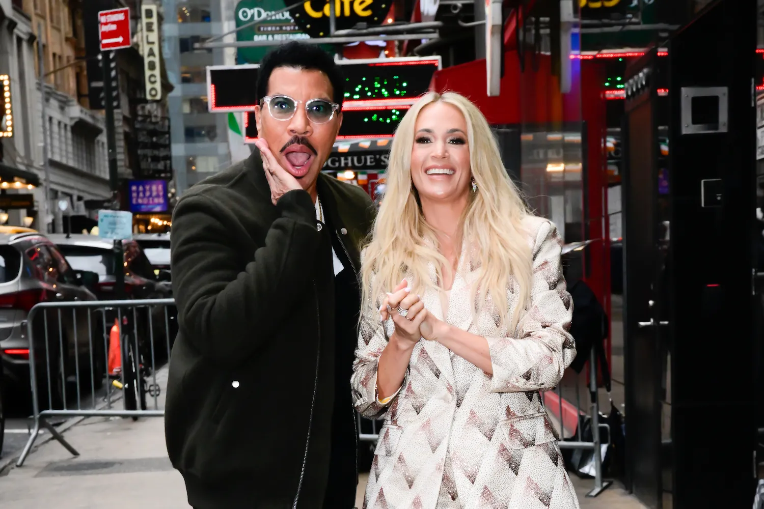 'American Idol' Season 23 judges Lionel Richie and Carrie Underwood posing in New York City outside of 'GMA.' Richie is posing with his mouth open and a hand to his cheek while Underwood laughs.