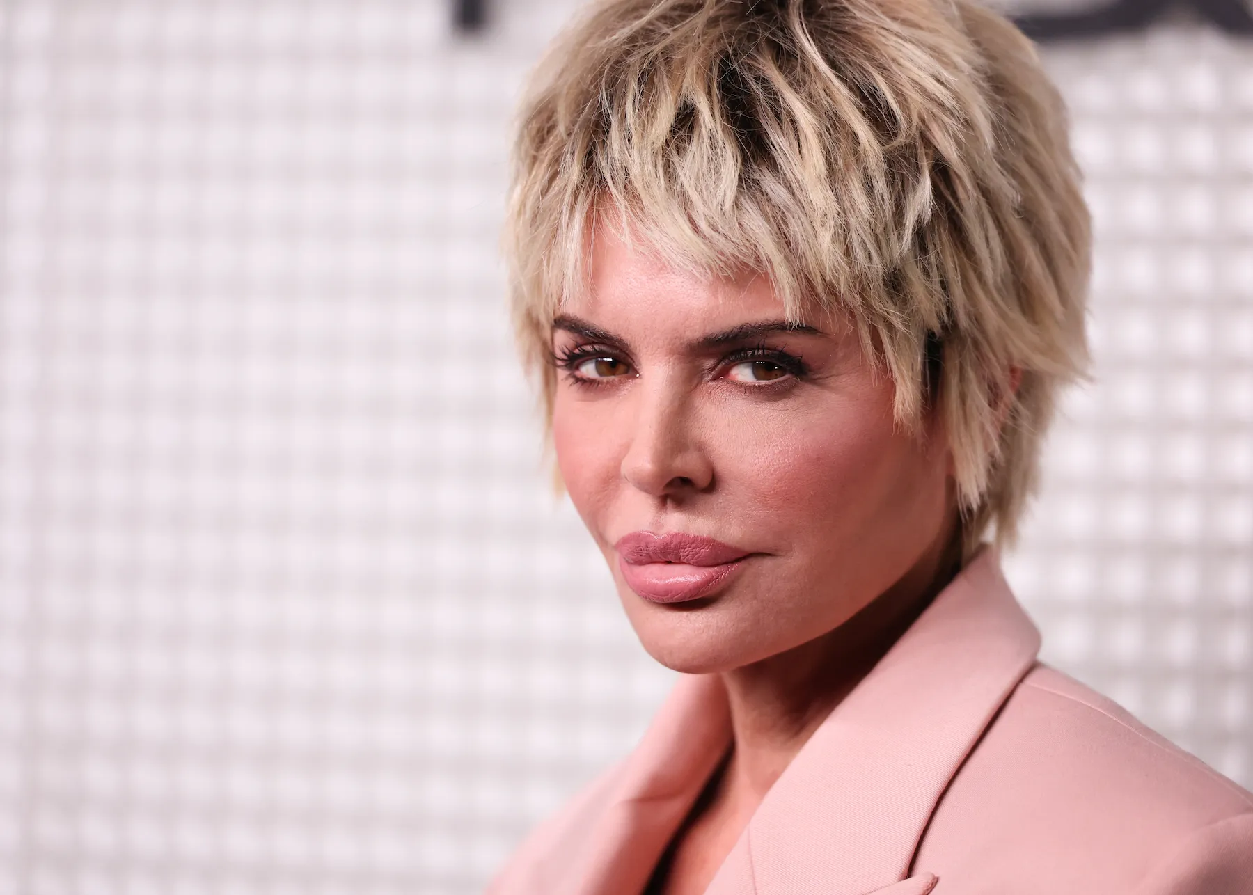 A close-up of 'The Real Housewives of Beverly Hills' star Lisa Rinna at an event. she's wearing a light pink suit and is against a pink background.