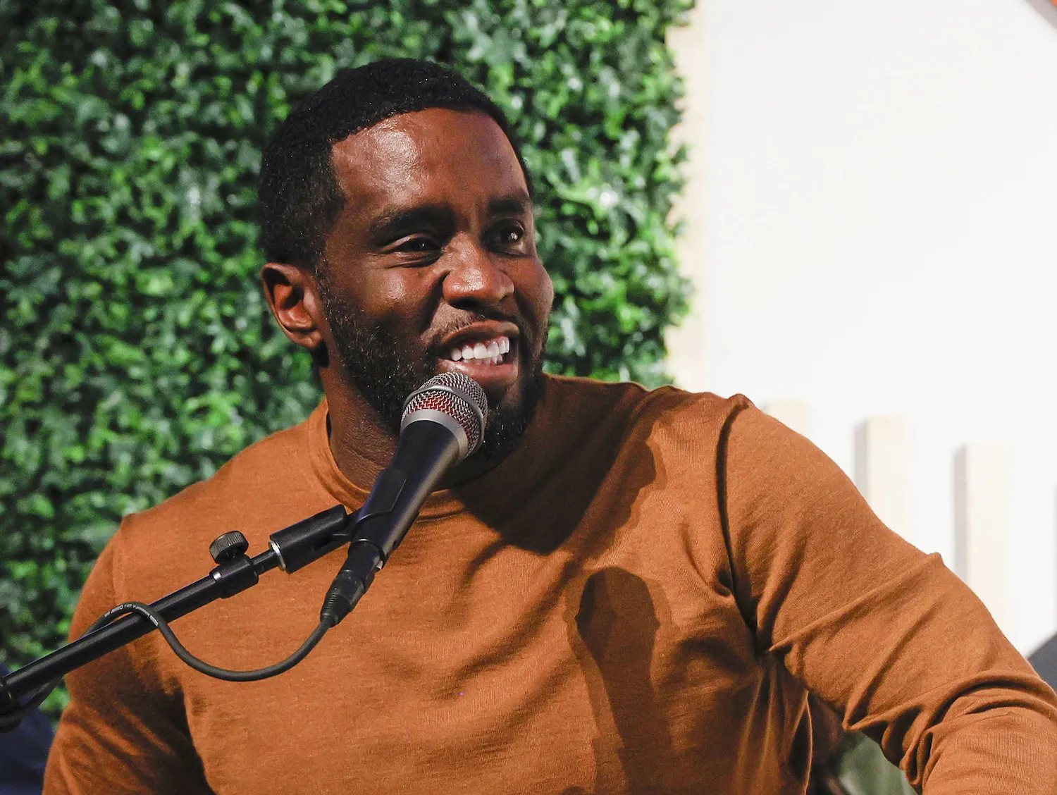 Sean 'Diddy' Combs smiling on a podium while speaking into a microphone. He's wearing an orange-brown sweater.