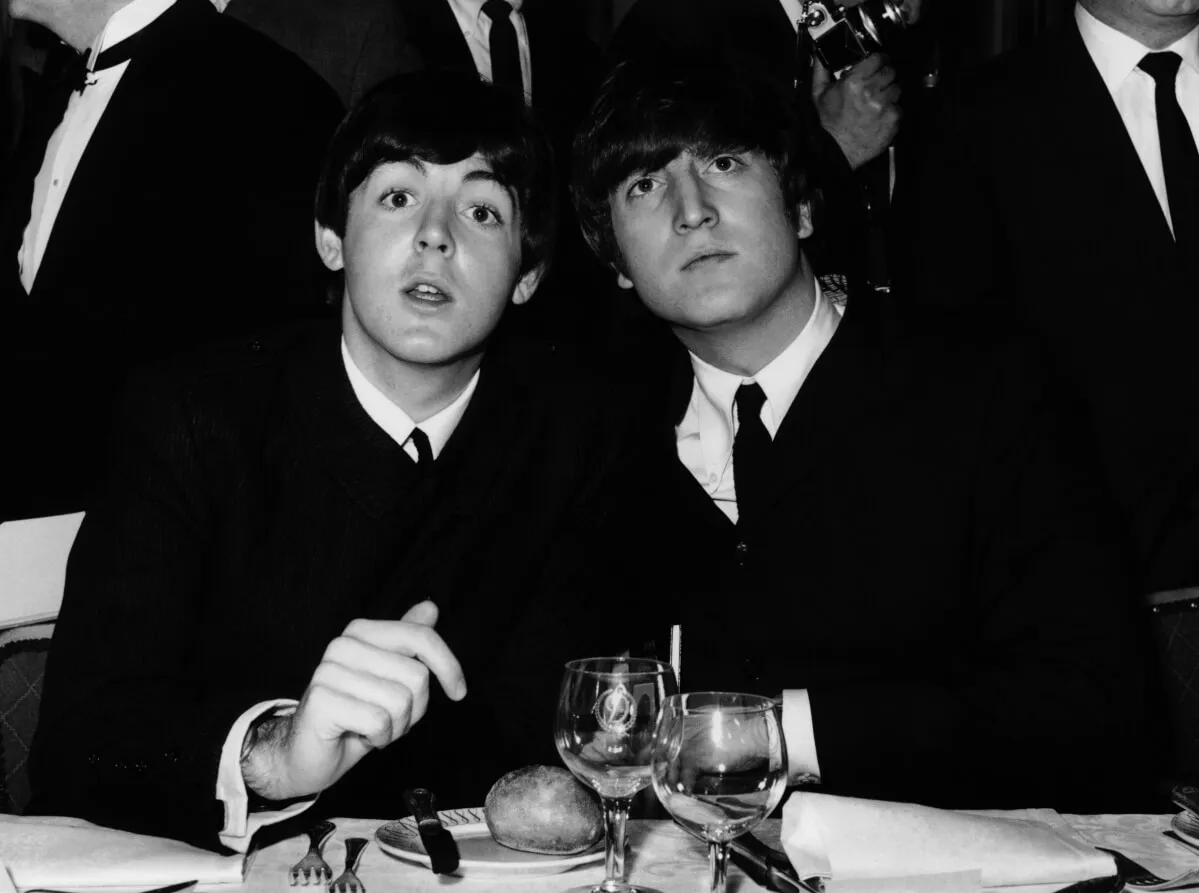 A black and white picture of Paul McCartney and John Lennon sitting at a table together. They wear suits.